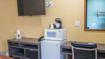 A hotel room with a desk, a microwave, a coffee maker, a TV mounted on the wall, and a chair.