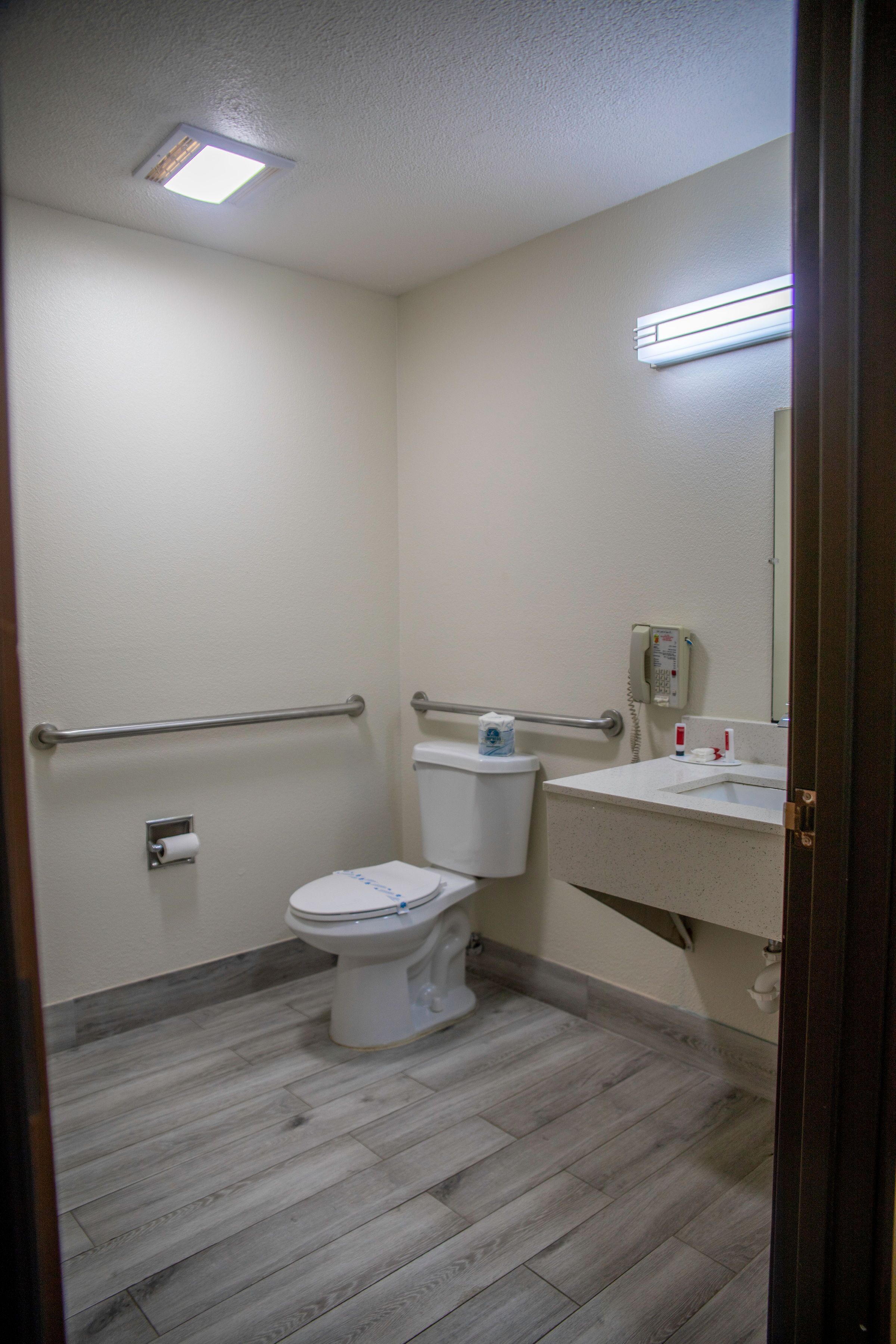 A small, clean bathroom with a toilet, a sink, a mirror, and a handrail.