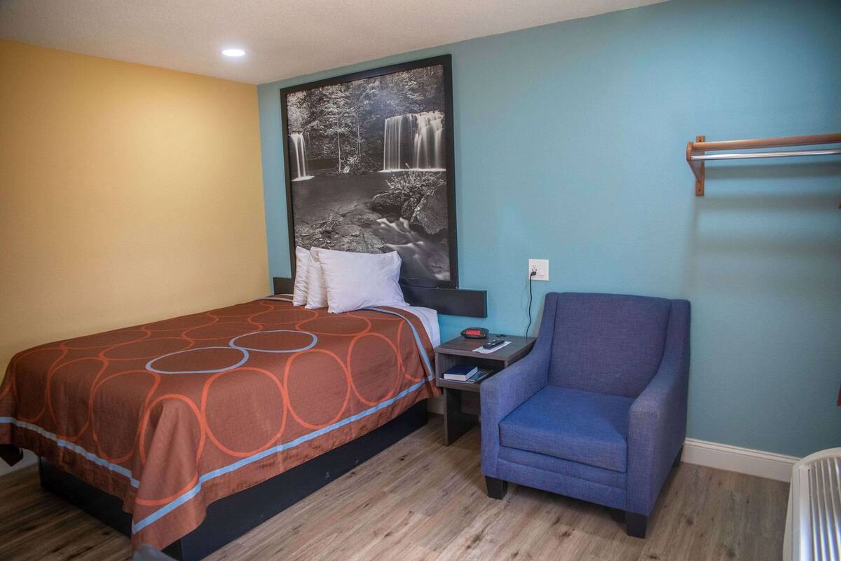 A hotel room with a bed, a chair, a nightstand, a wall-mounted TV, and a power outlet.