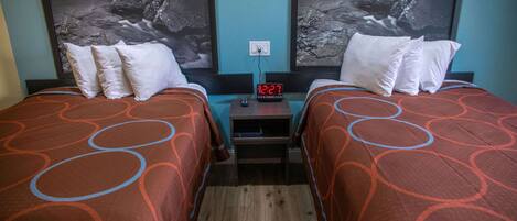 A hotel room with two beds, each with a patterned bedspread, a nightstand with a digital clock, and a wall-mounted outlet.