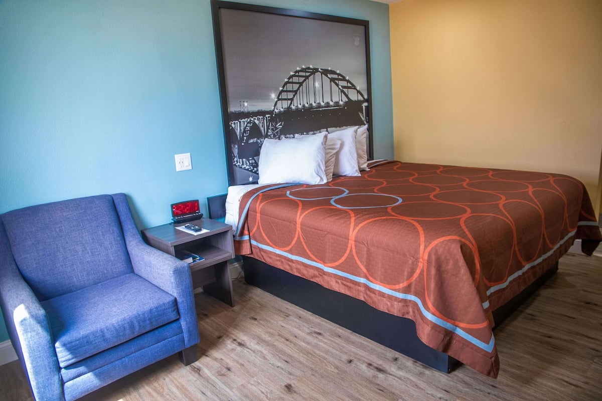 A hotel room with a bed, a chair, a nightstand, and a wall art of a bridge.