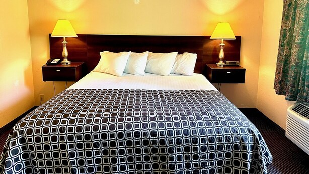 A hotel room with a large bed, two bedside tables with lamps, a headboard, and a window with curtains. - Haven Inn & Suites (Monroe)