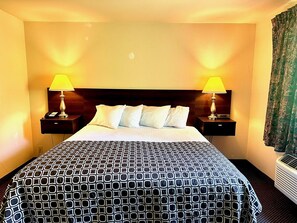 A hotel room with a large bed, two bedside tables with lamps, a headboard, and a window with curtains. - Haven Inn & Suites (Monroe)