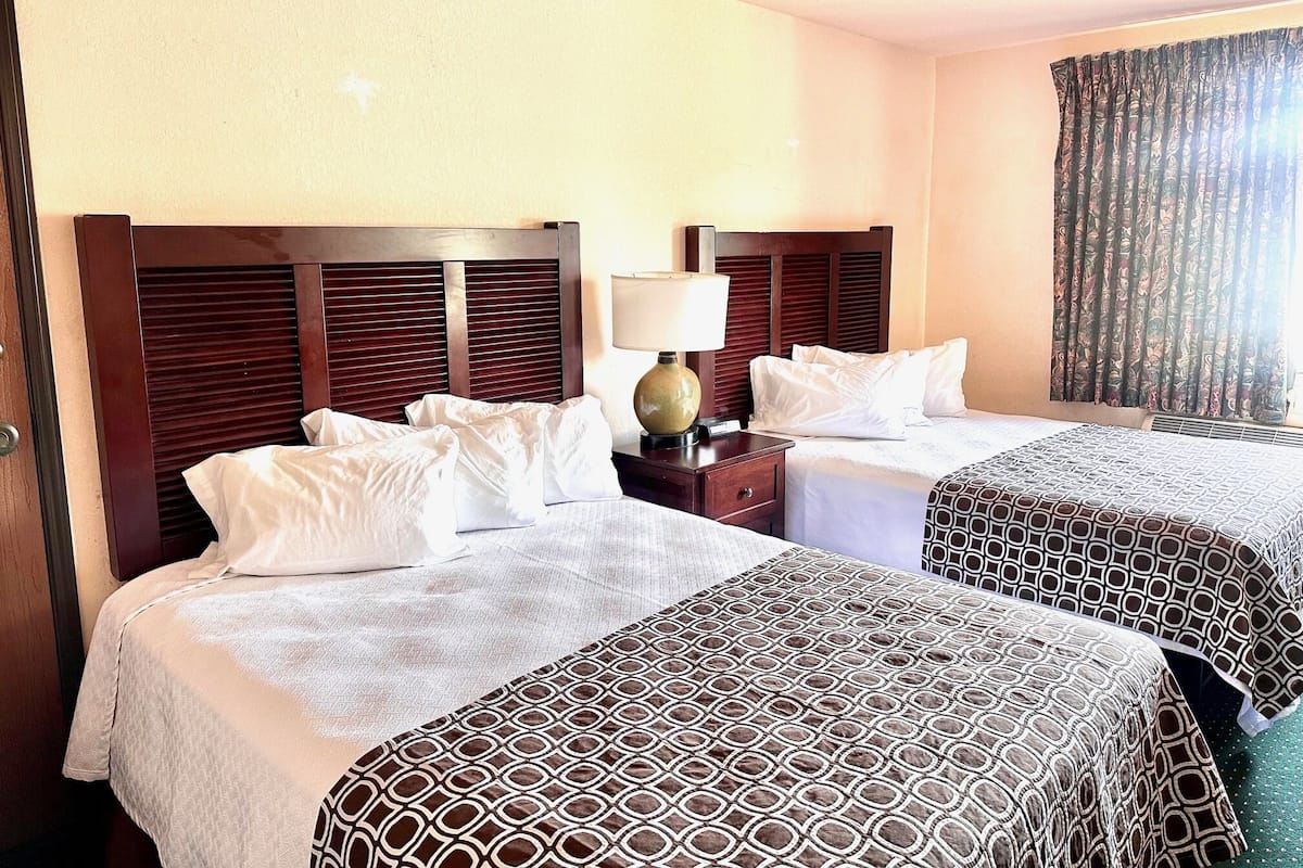 A hotel room with two beds, a nightstand with a lamp, and a window with curtains.