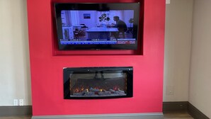 A flat-screen TV mounted on a red wall, displaying financial news. Below it, a fireplace with logs is visible. - Haven Inn & Suites (Monroe)