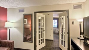 Quarto de hotel com piso de carpete, uma cadeira, escrivaninha com luminária e televisão.