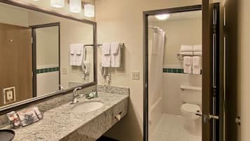 Un baño con encimera de granito, un lavamanos, un espejo y zona de ducha con puerta de vidrio.