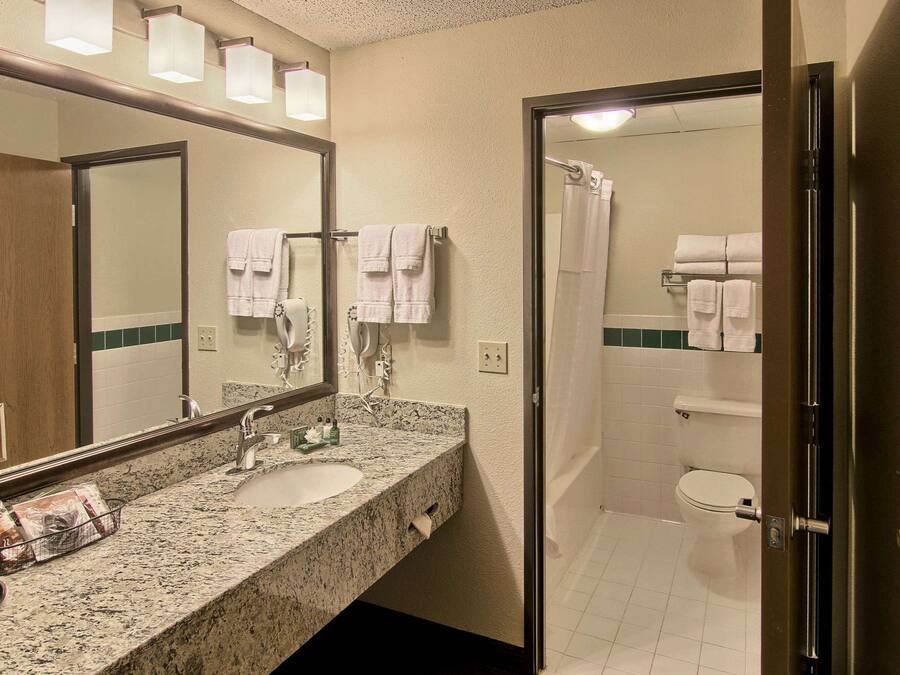Un baño con encimera de granito, un lavamanos, un espejo y zona de ducha con puerta de vidrio.