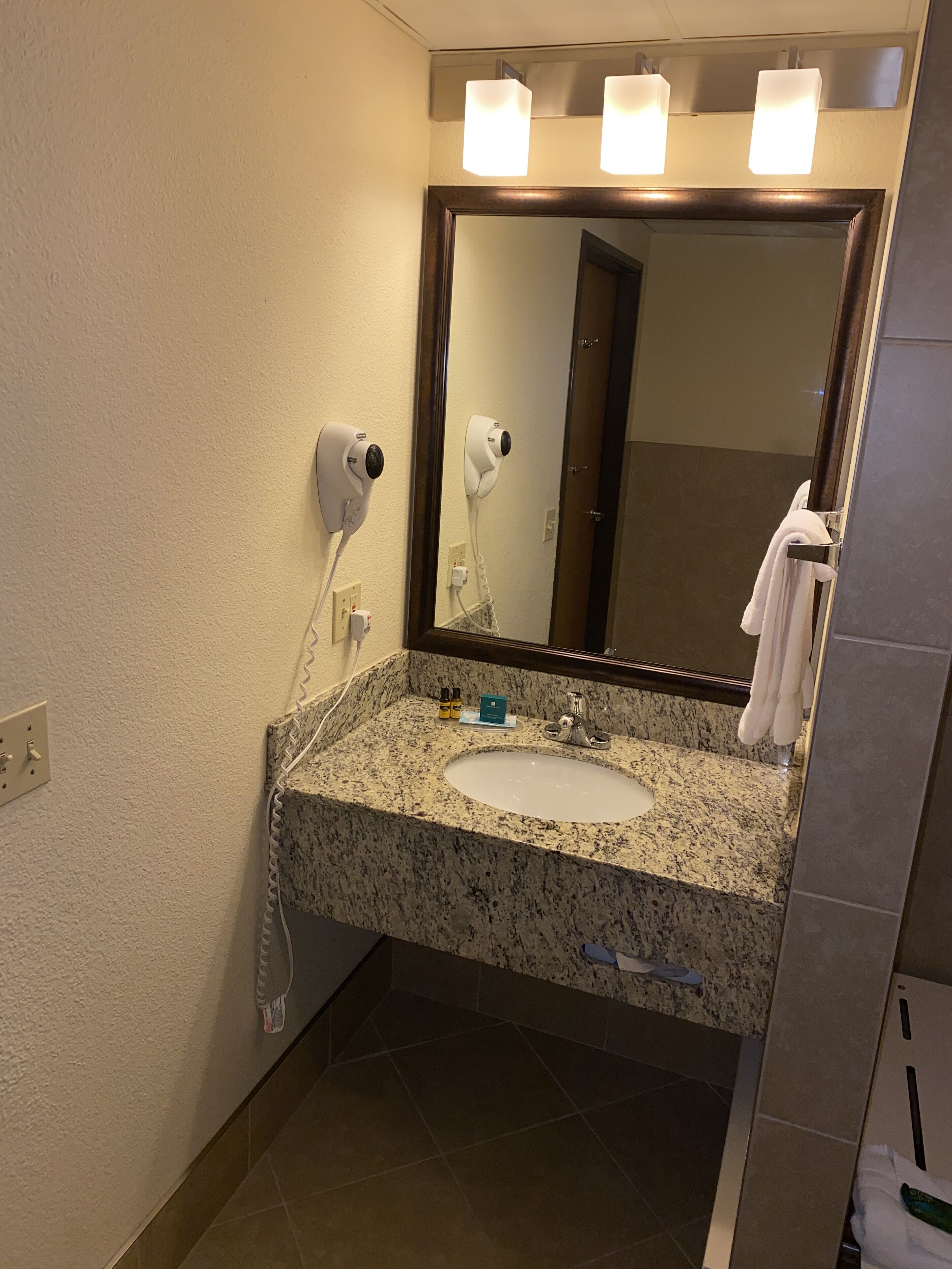 A bathroom with a granite countertop, a white sink, a mirror with lights, a hairdryer, and a towel rack.