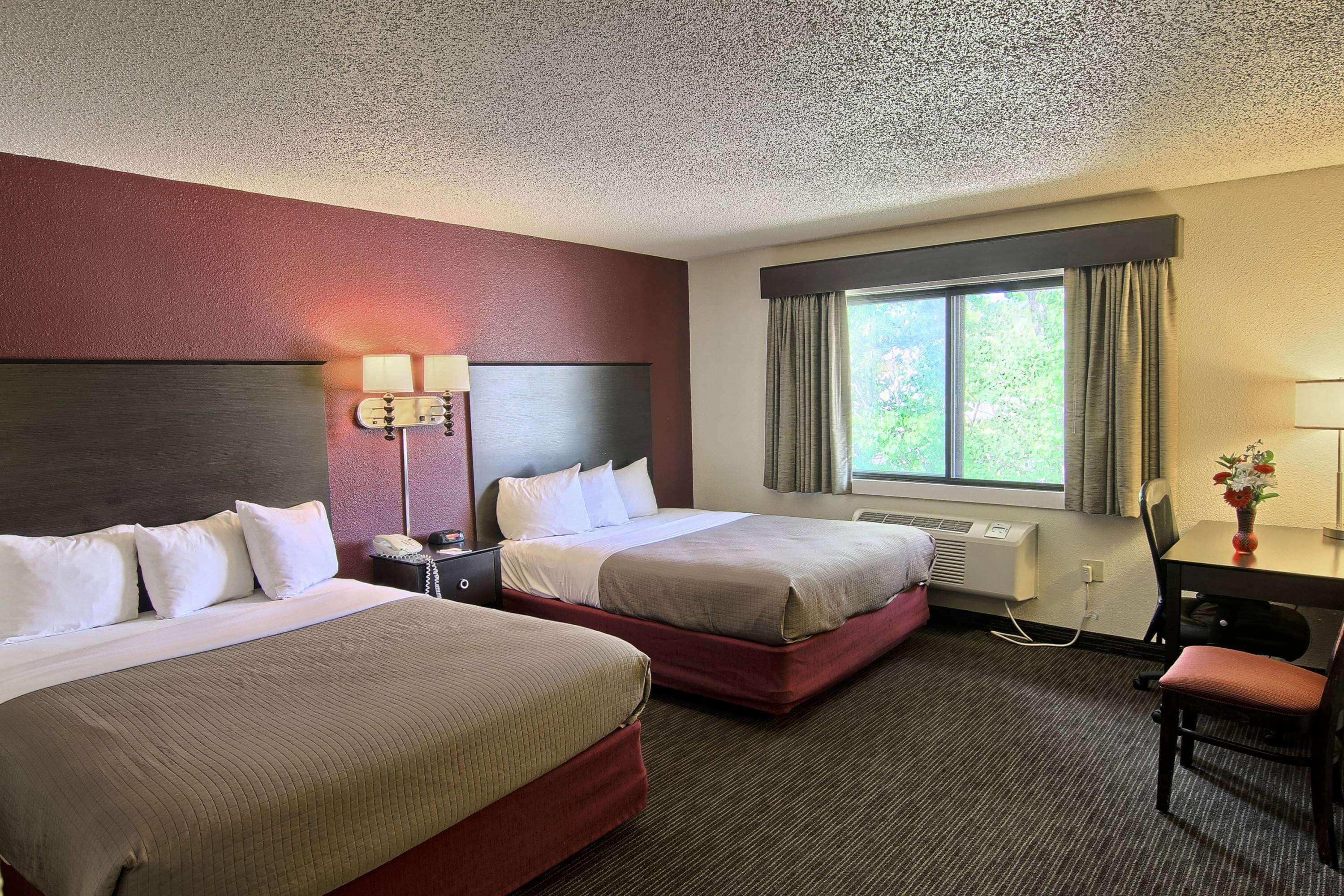 A hotel room with two beds, a desk, a chair, a lamp, and a window with curtains.