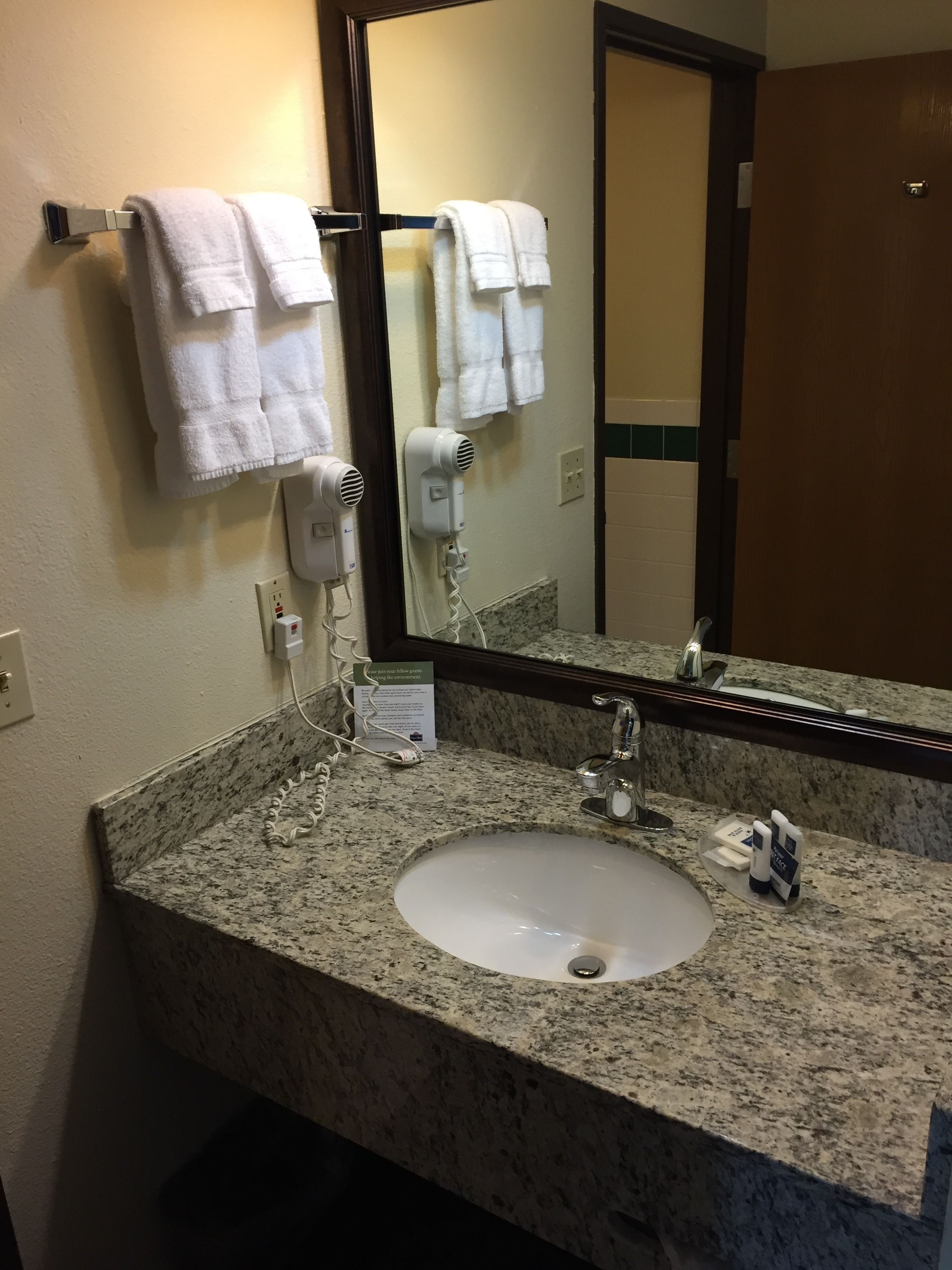 A hotel bathroom with a granite countertop sink, a mirror, two hair dryers, and a towel rack with multiple towels.