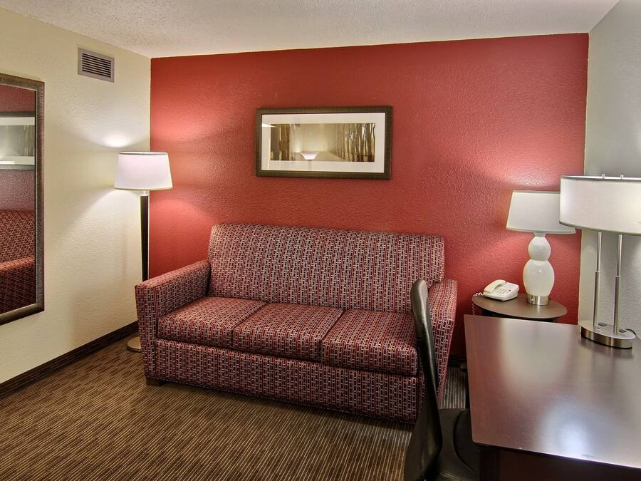 Quarto de hotel com parede vermelha, sofá estampado, espelho, mesa de cabeceira com abajur e quadro emoldurado.