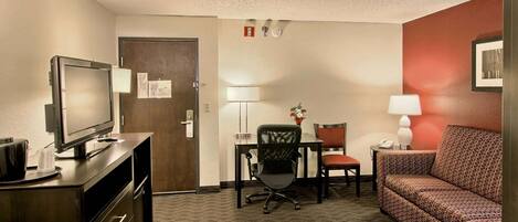 A hotel room with a television, desk, chair, sofa, and a door.