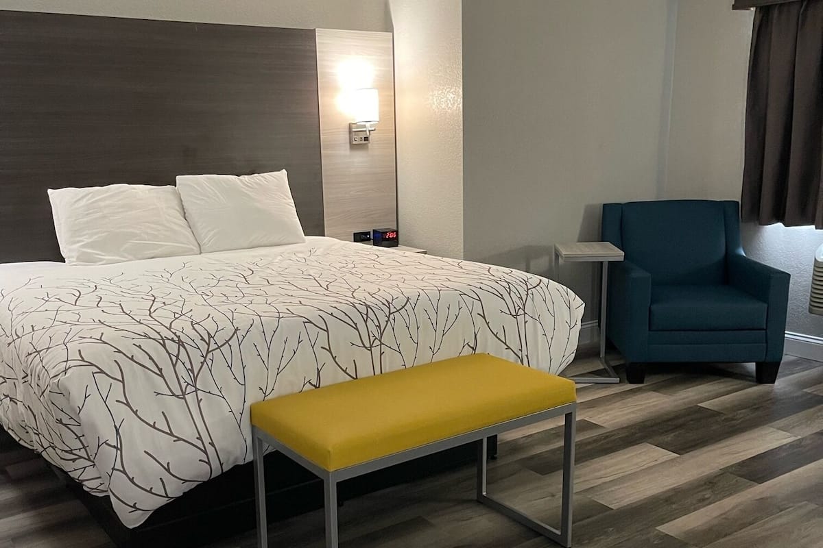 A hotel room with a large bed, a bedside table, a chair, and a bench.