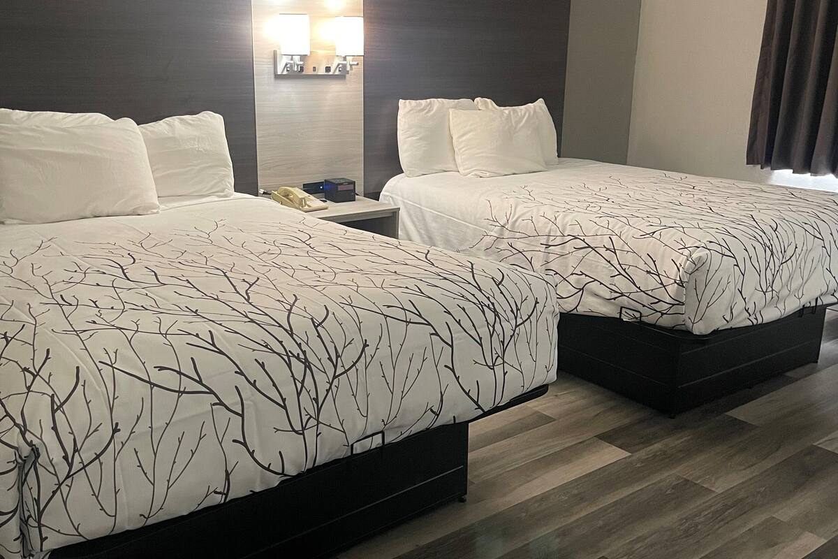 A hotel room with two beds, each with a dark headboard and white bedding. There is a nightstand with a lamp between the beds. The room has a wooden floor and a large window with curtains.