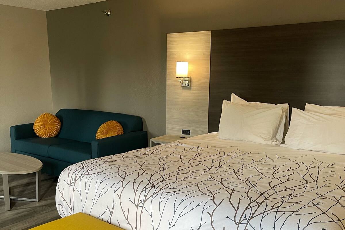 A hotel room with a large bed, a small table, a bench, a sofa, and a wall-mounted light.