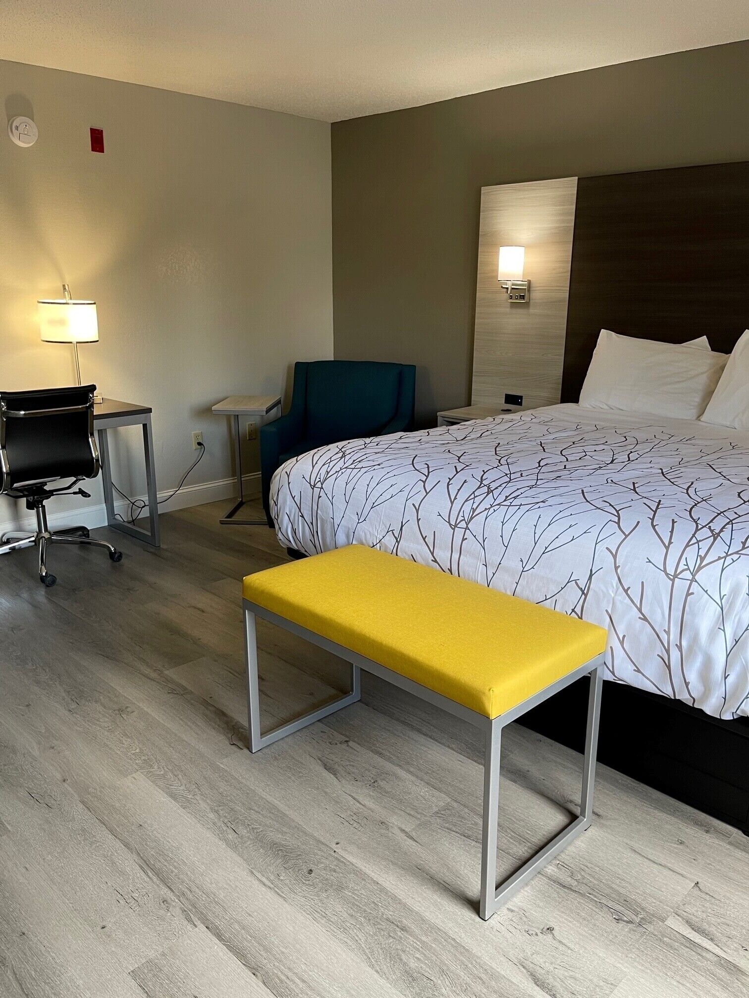 A hotel room with a bed, a desk, a chair, a bench, and a wall-mounted lamp.
