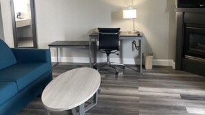 A modern hotel room with a blue sofa, a small round table, a desk with a chair, a lamp, a TV, and a fireplace. - HomeTown Inn & Suites (Belle Plaine)