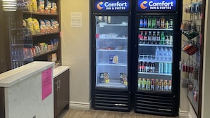 Zona de recepción de un hotel con dos máquinas expendedoras identificadas como 'Comfort Inn & Suites', un mostrador con una nota rosa y estantes llenos de aperitivos y agua embotellada.