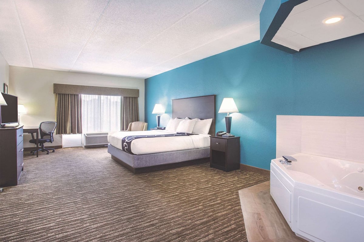 Habitación de hotel con cama, lámparas de noche, escritorio con silla, lámpara de escritorio, ventana grande con cortinas y bañera.