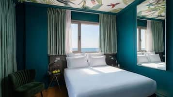 Habitación de hotel con una cama grande, una silla, un escritorio con botellas y vistas al mar a través de una ventana.