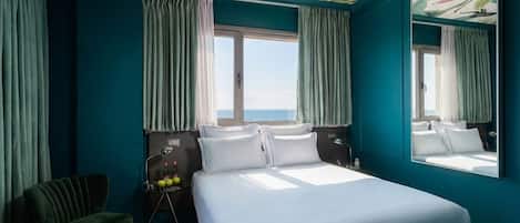 A hotel room with a large bed, a chair, a desk with bottles, and a view of the sea through a window.
