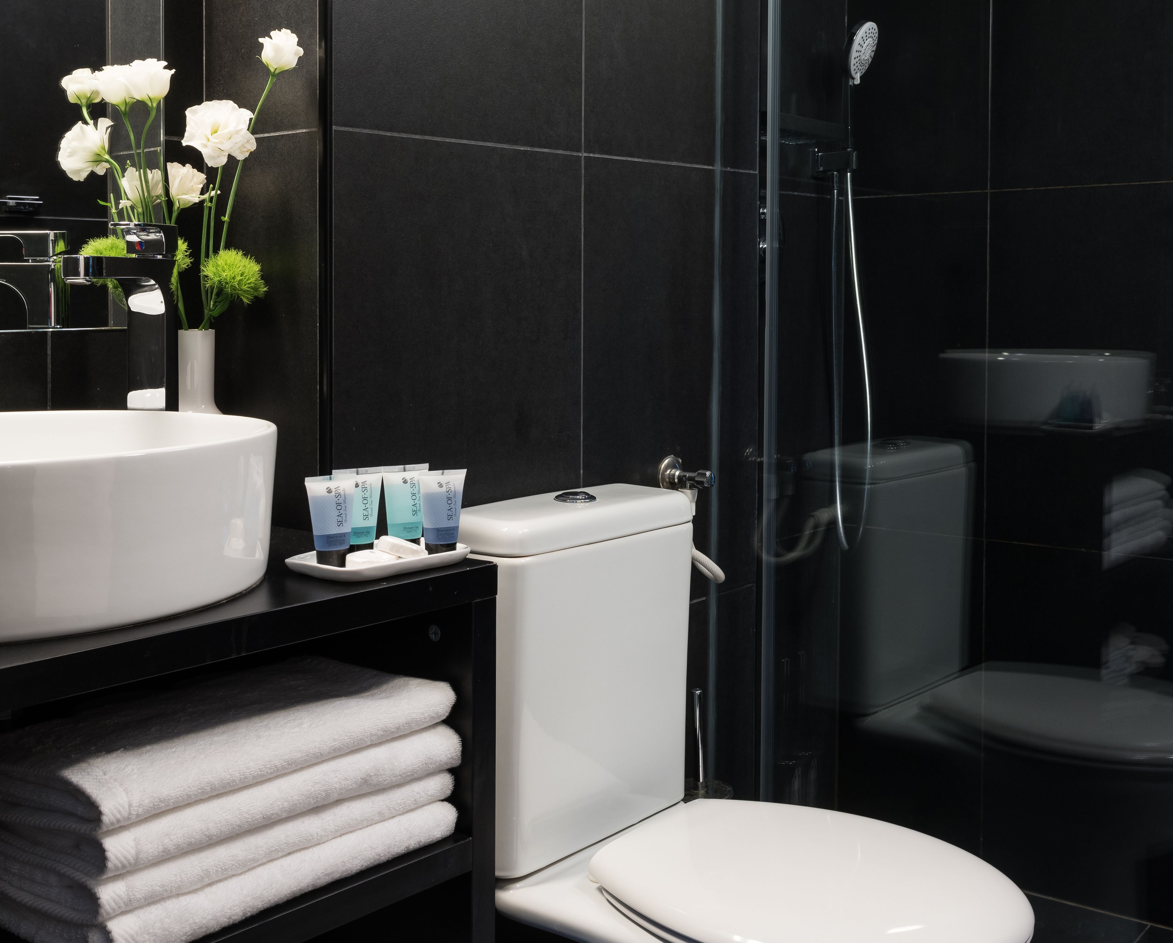 A modern bathroom with a white sink, toilet, and shower. There are towels stacked on a shelf and a vase with white flowers.