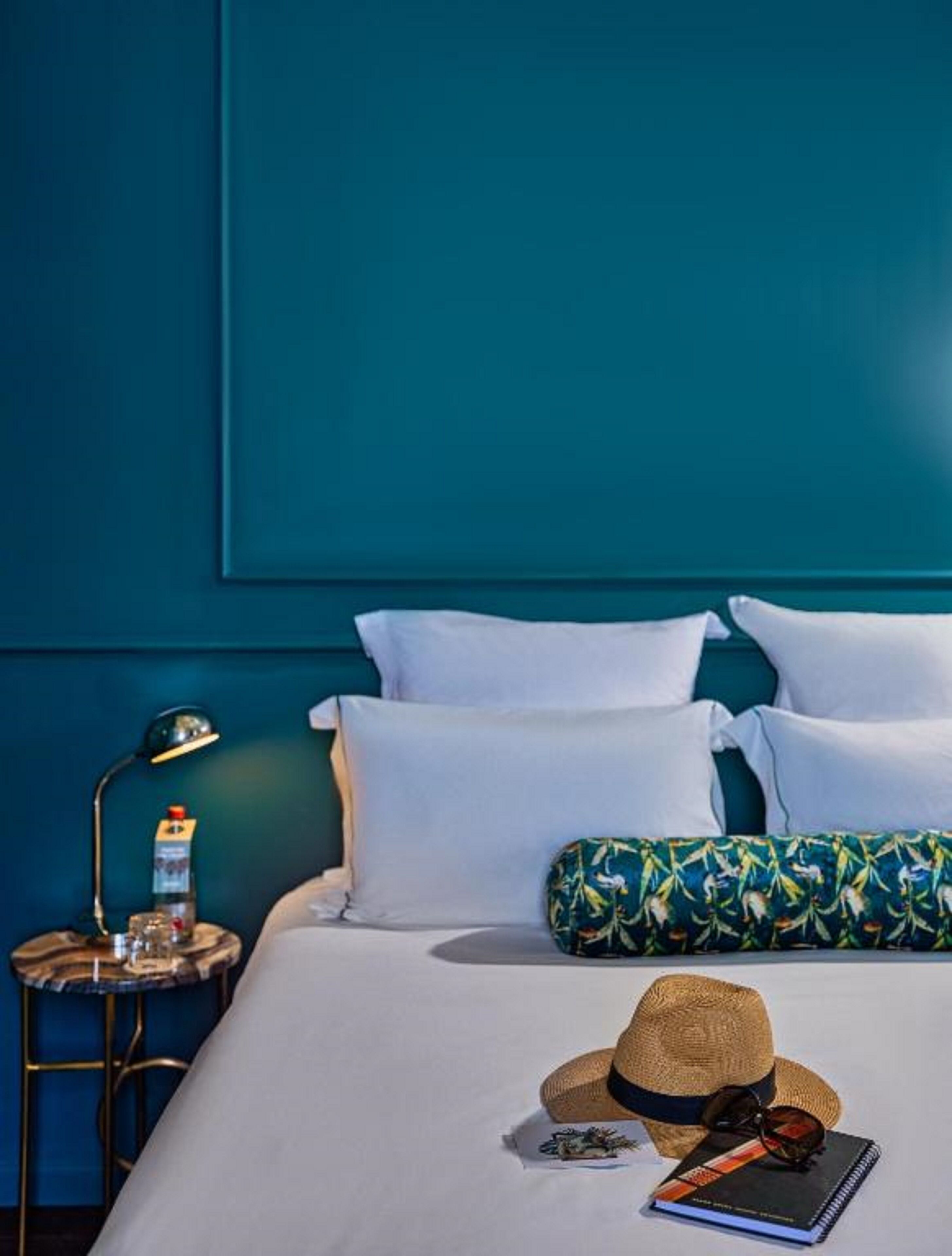Een bed met witte lakens, een strohoed, zonnebril en een notitieboekje tegen een wand in teal.