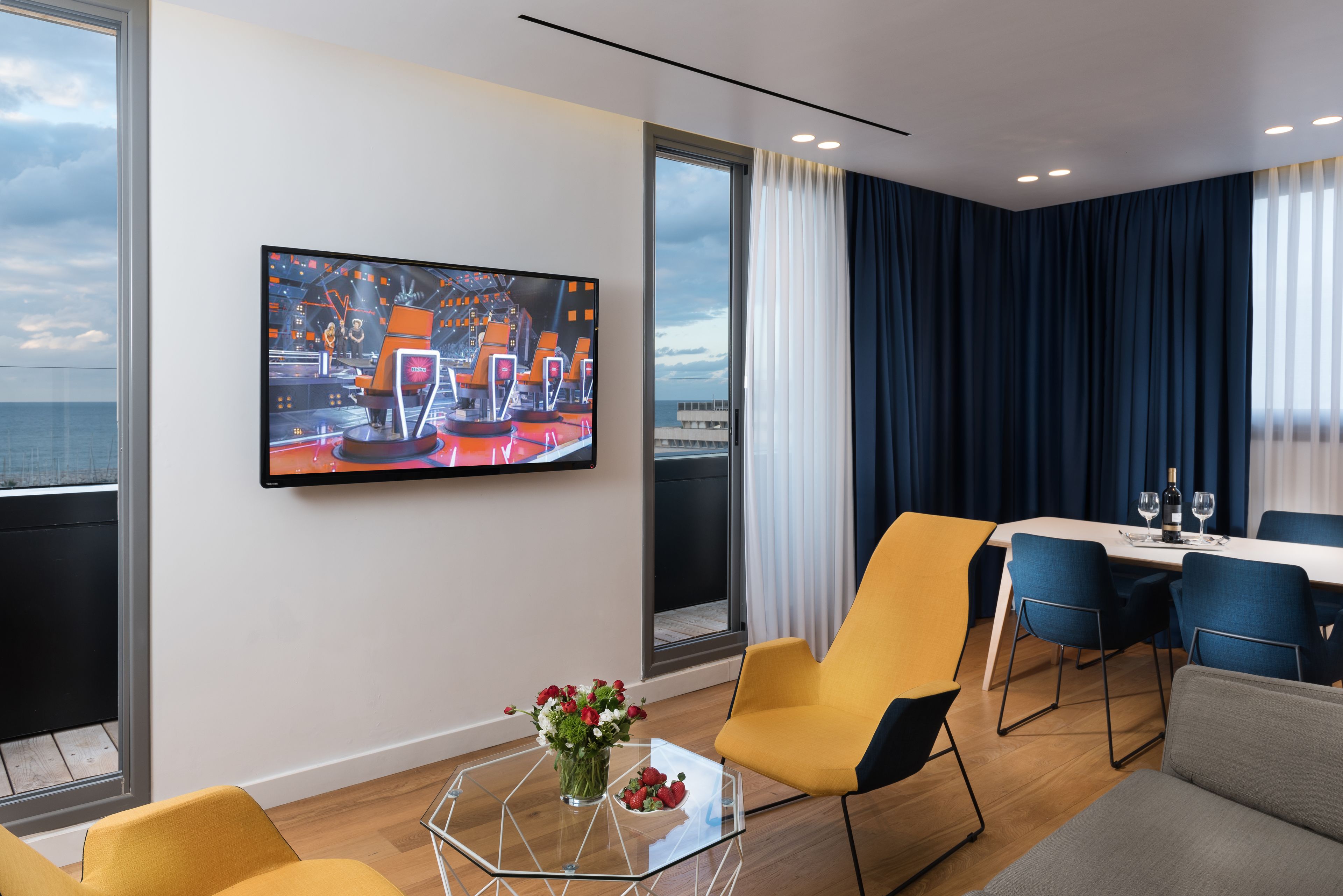 A modern hotel room with a large TV, a dining area, and a view of the sea.