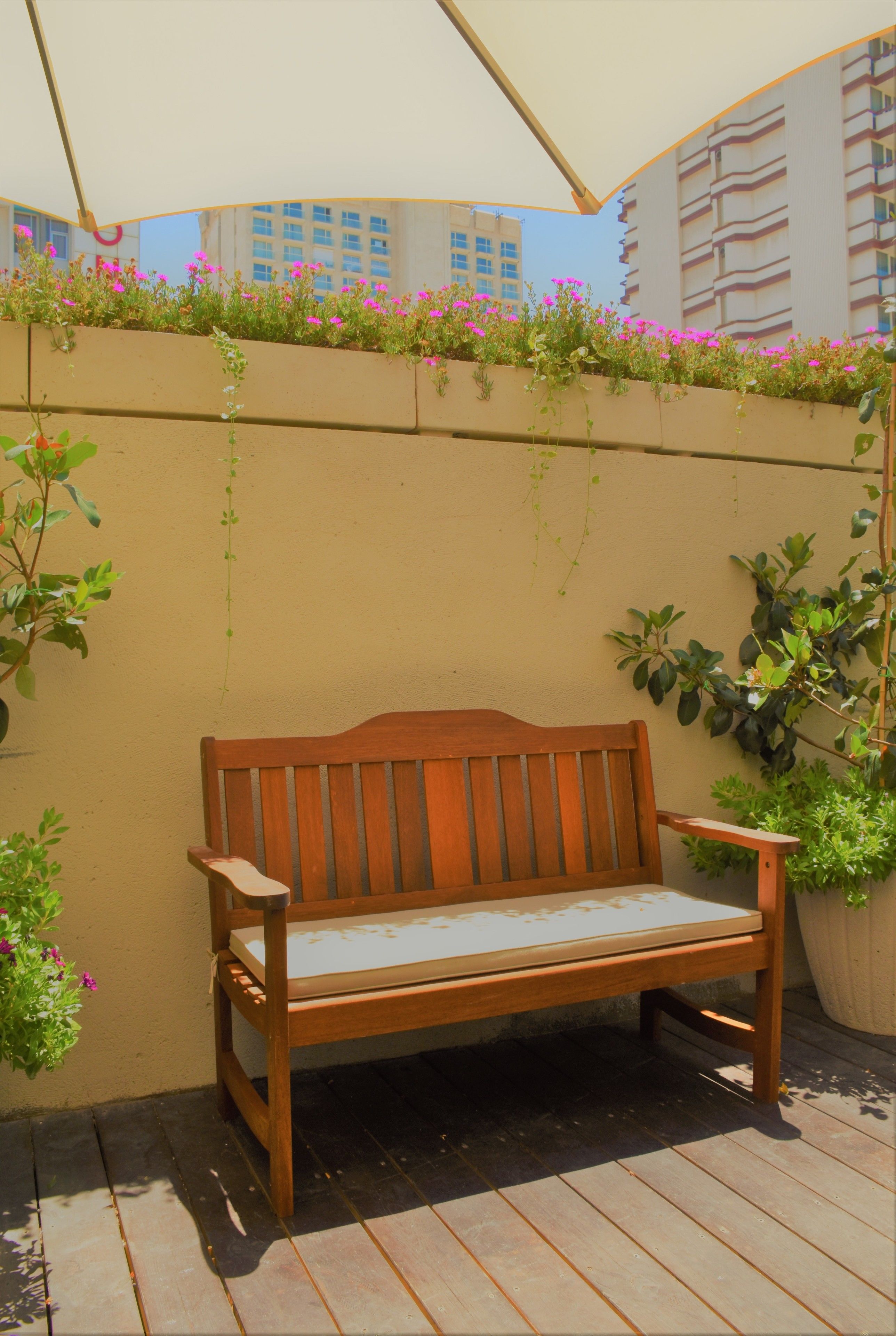 Un banc en bois avec un coussin, placé sur une terrasse en bois, avec un mur beige en arrière-plan.