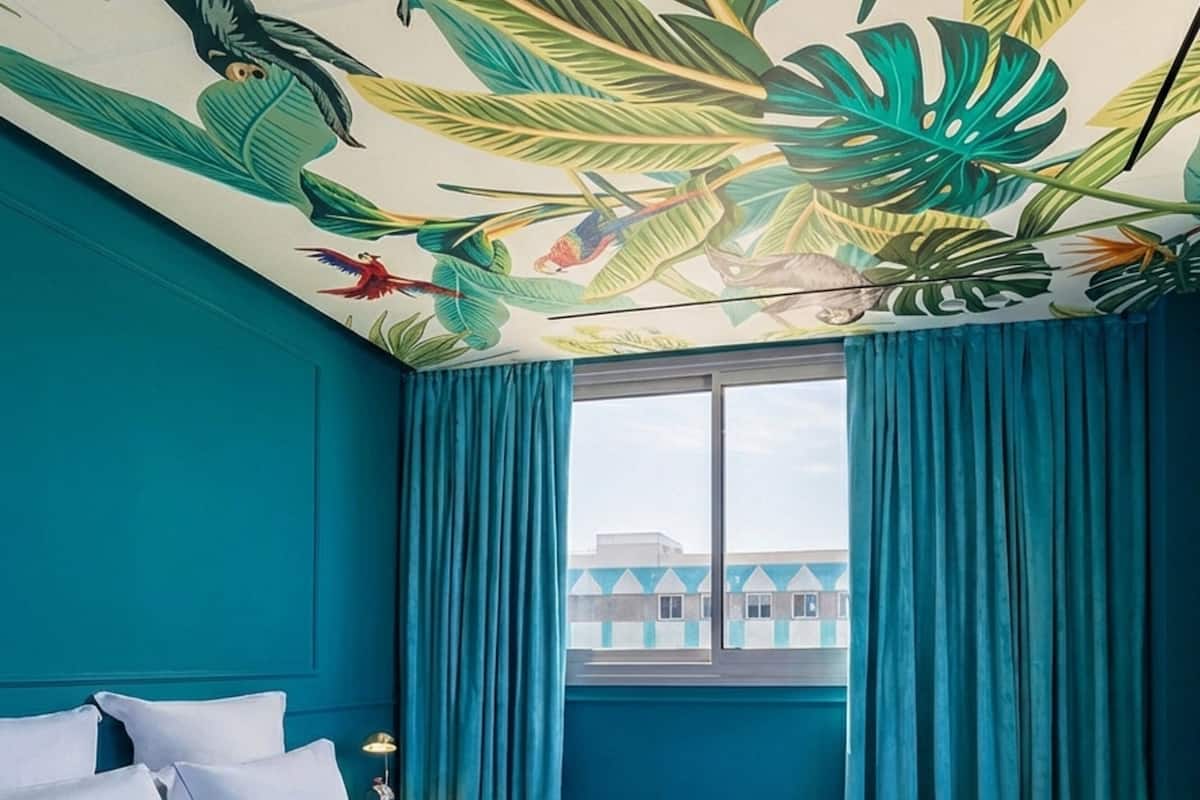Une chambre d’hôtel avec un grand lit, des murs de couleur turquoise, une fresque murale aux motifs tropicaux et une vue sur les bâtiments par la fenêtre.