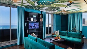 Ein modernes Wohnzimmer mit türkisfarbenen Sofas, einem großen Flachbildfernseher und einem Wandgemälde mit tropischen Blättern an der Decke.