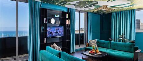 Un salón moderno con sofás color turquesa, un televisor de pantalla plana grande y un mural en el techo con hojas tropicales.