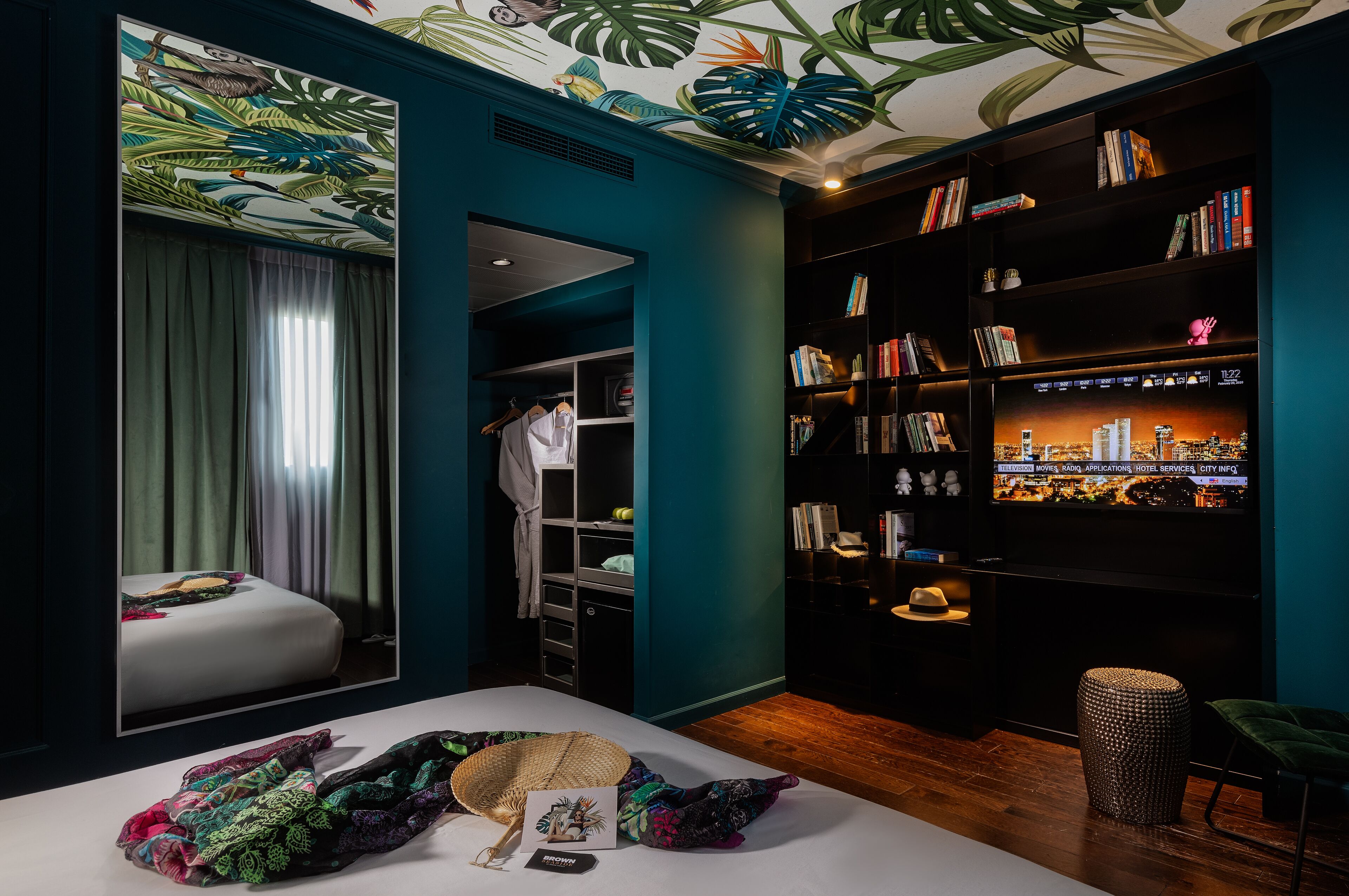 Ein Hotelzimmer mit einem Bett, einem großen Spiegel, einem Bücherregal und einem Fernseher, der eine Stadtansicht zeigt.