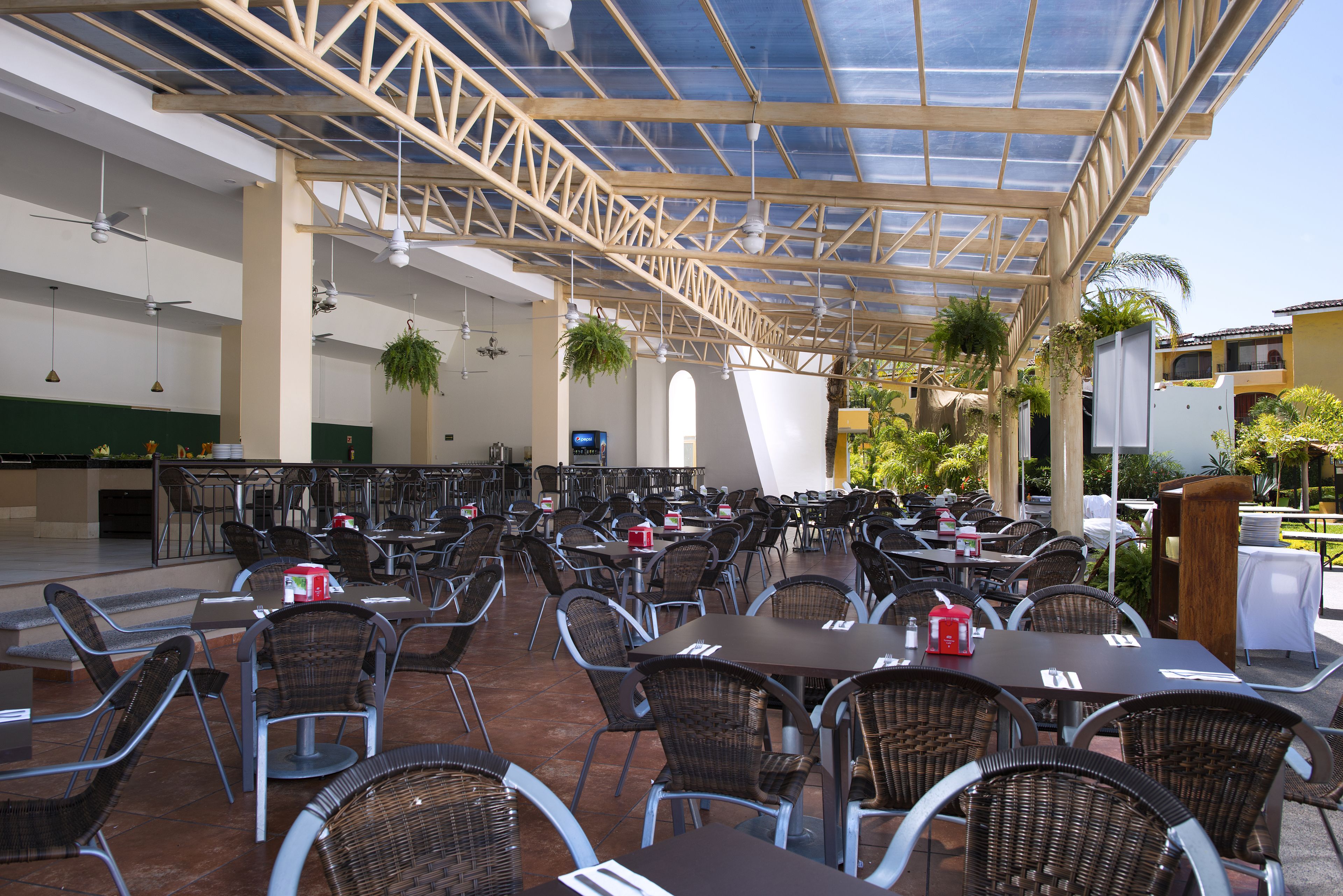 Un espace extérieur abrité, aménagé pour les repas, avec plusieurs tables et chaises, une structure au plafond et des plantes suspendues.