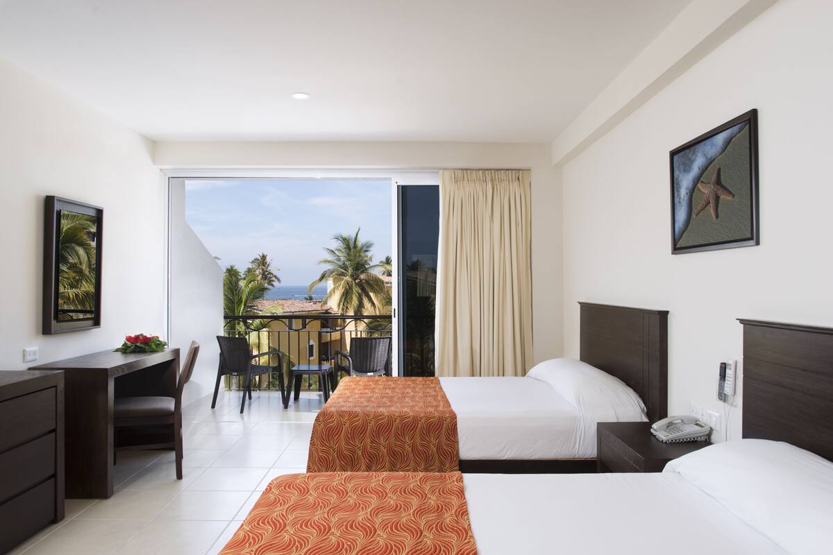 A hotel room with two beds, a desk, and a chair. There is a balcony with a view of palm trees and the ocean.