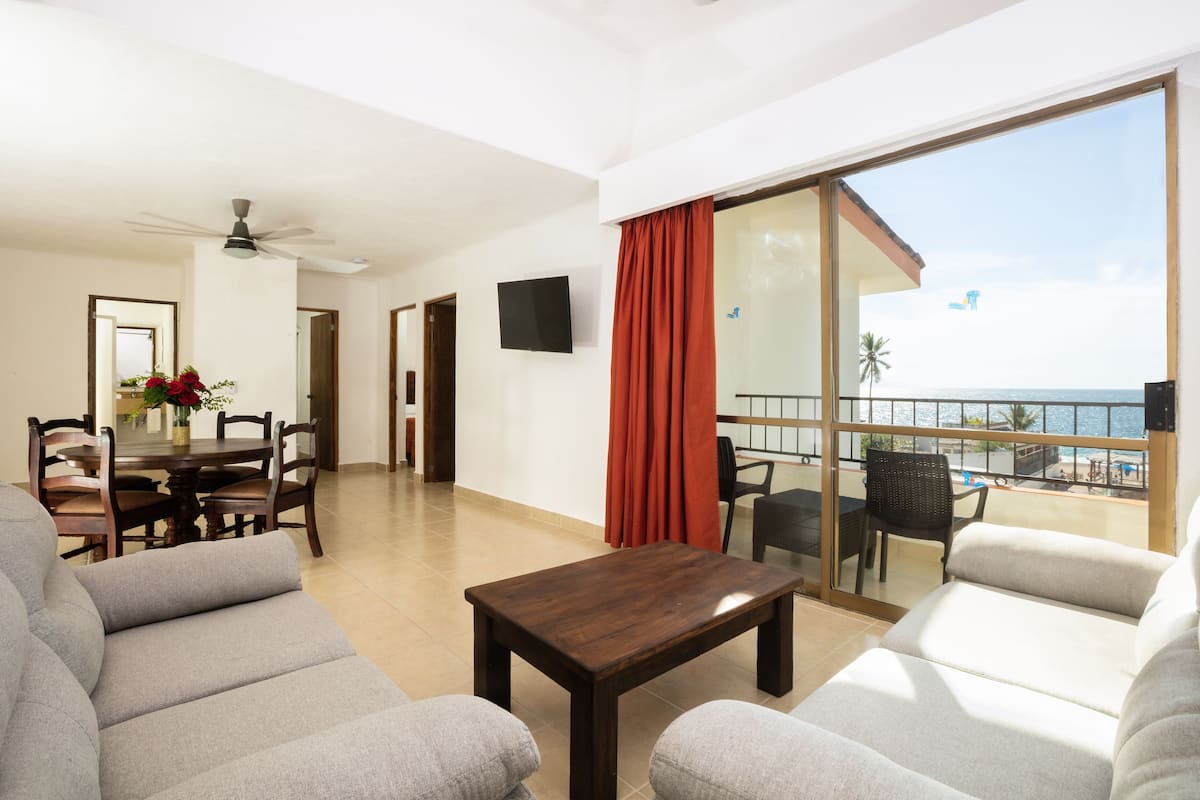 A spacious living area with a balcony overlooking the sea, a dining table with chairs, and a flat-screen TV mounted on the wall.