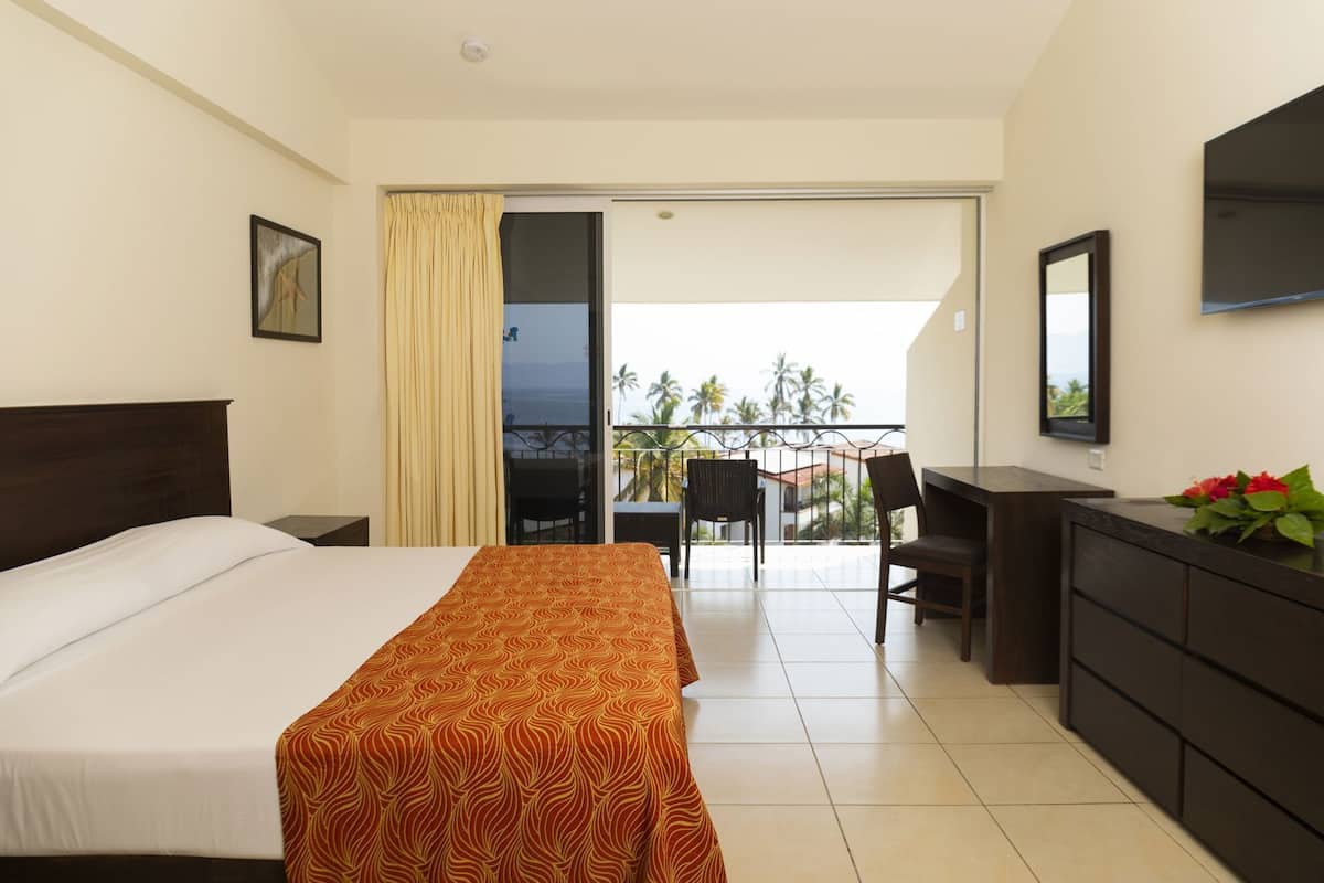 A hotel room with a bed, a desk, a dresser, a balcony with a view of palm trees, and a sliding door.
