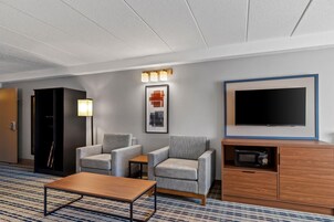 Habitación de hotel con televisión de pantalla plana, dos sillones grises, una mesa de centro de madera, una mesita auxiliar pequeña, un cuadro enmarco, un microondas y un armario.