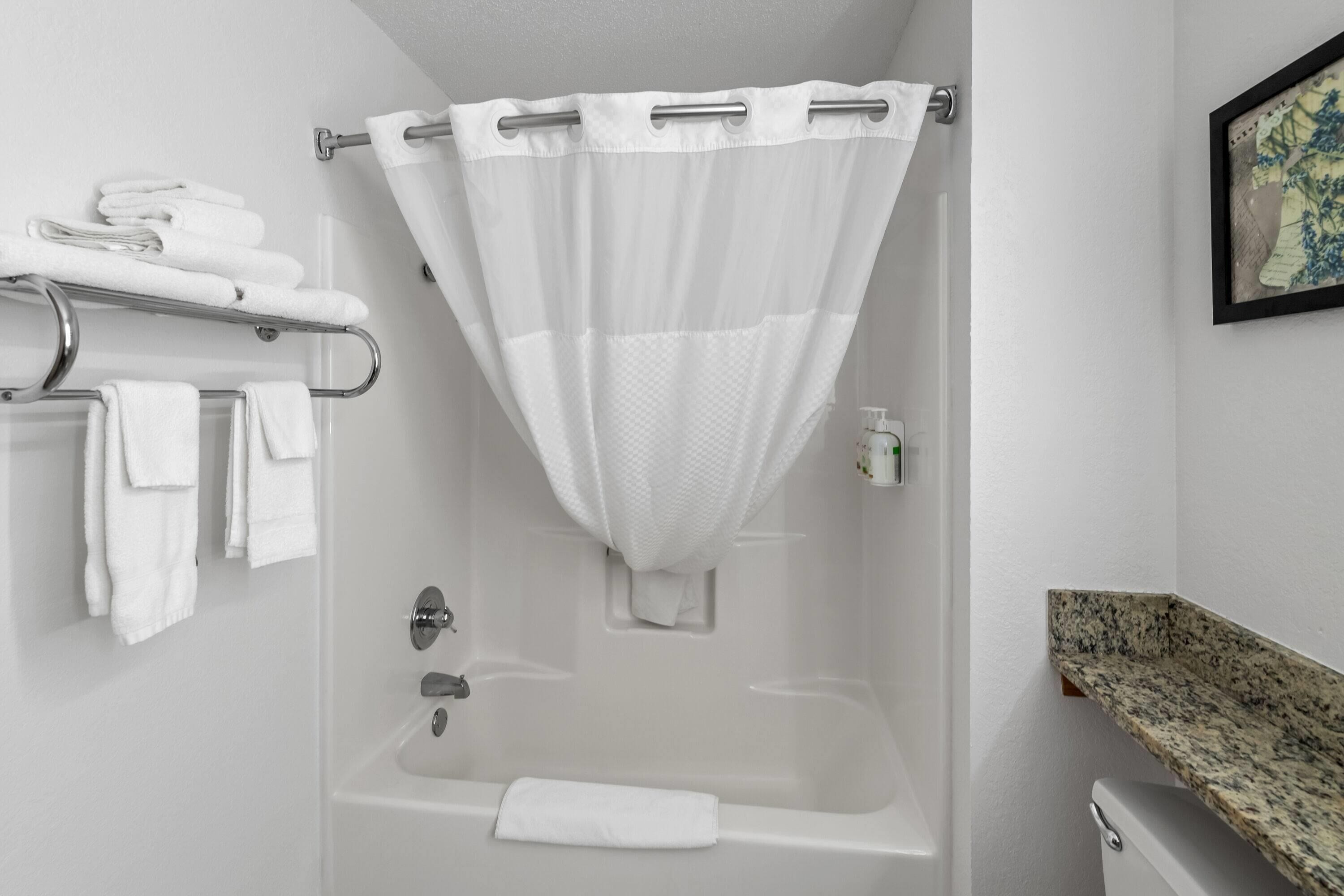 A bathroom with a shower, a towel rack with towels, a framed picture on the wall, and a granite countertop.