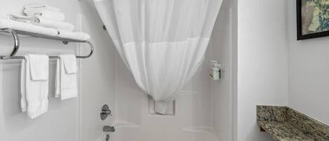 A bathroom with a shower, a towel rack with towels, a framed picture on the wall, and a granite countertop.