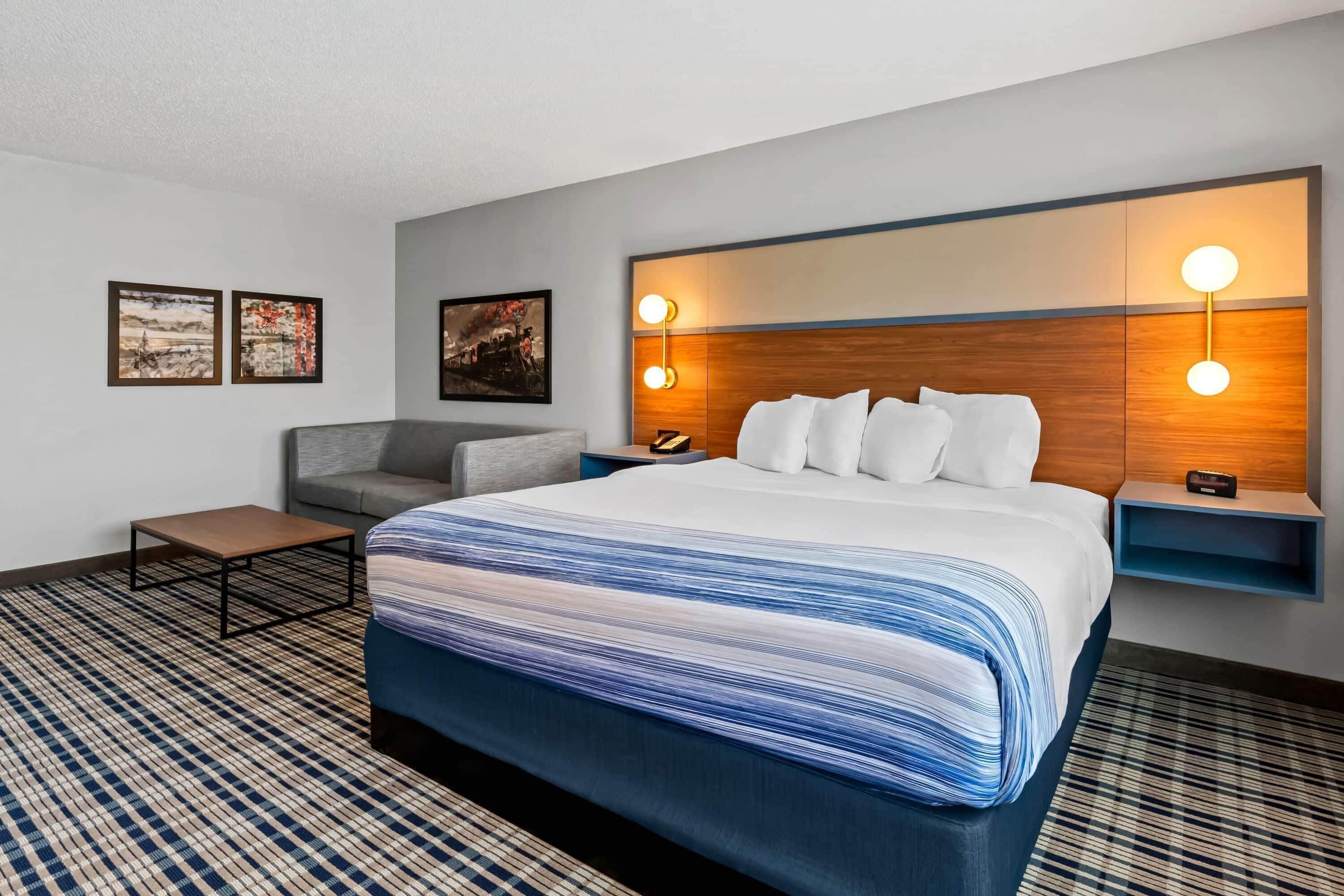 Quarto de hotel com uma cama grande, um sofá cinzento, uma mesinha e duas luminárias fixadas na parede.
