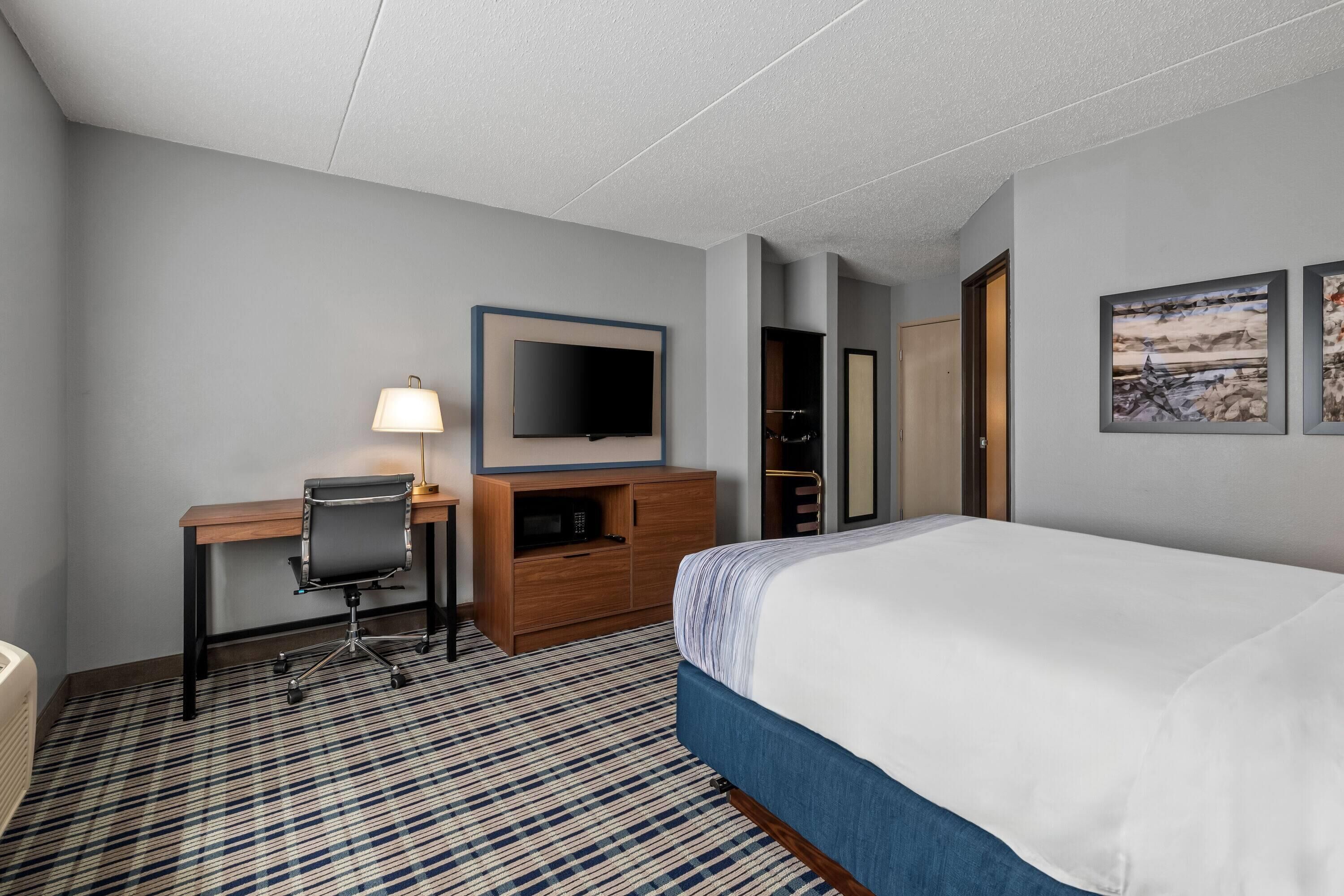 Ein Hotelzimmer mit Bett, Schreibtisch mit Stuhl, Fernseher, Kleiderschrank und einem gemusterten Teppich.