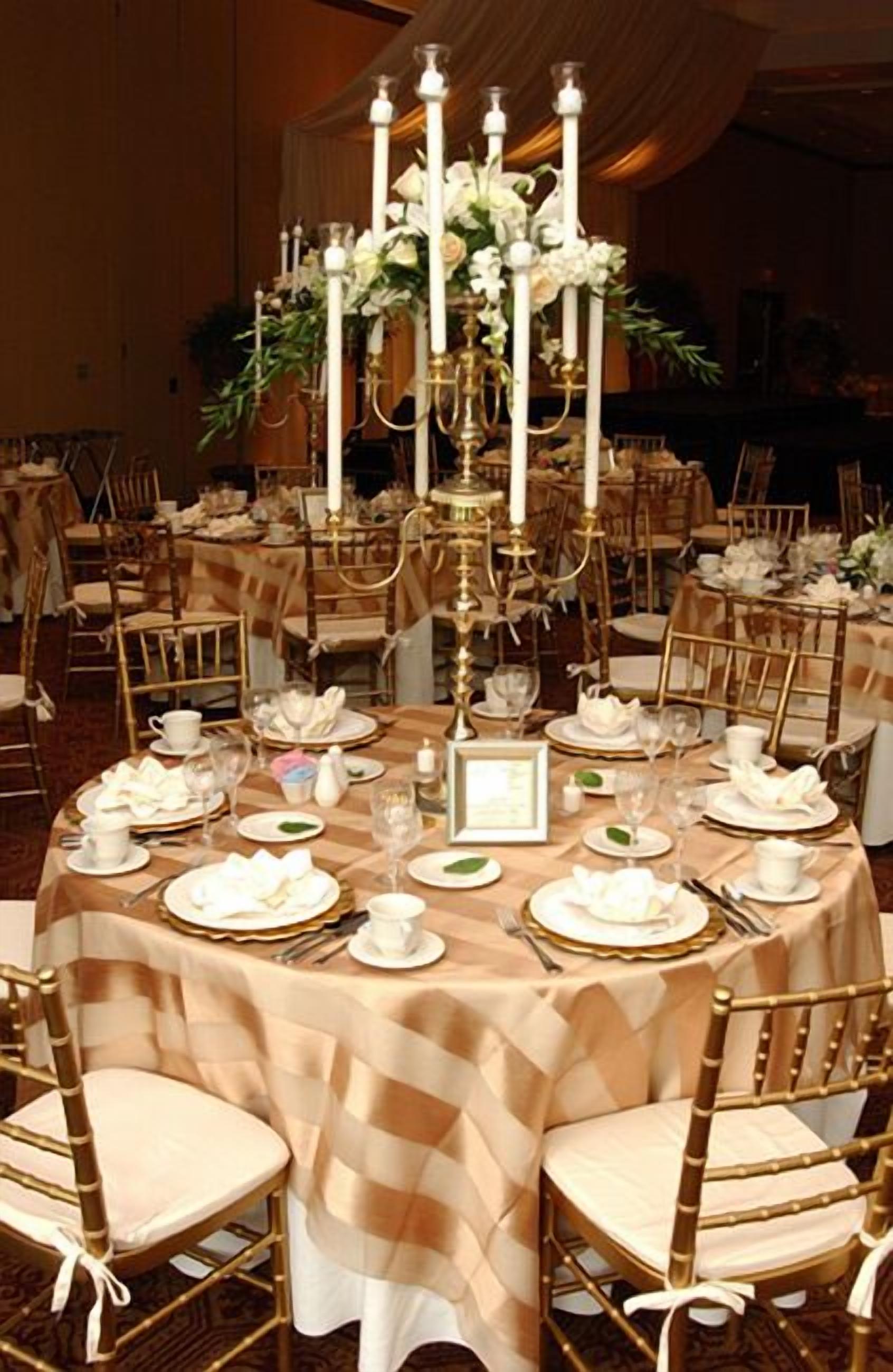 Ein Bankettsaal mit runden Tischen, gedeckt für eine festliche Veranstaltung, mit goldenen Stühlen, weißen Tischdecken und floralen Mittelstücken.