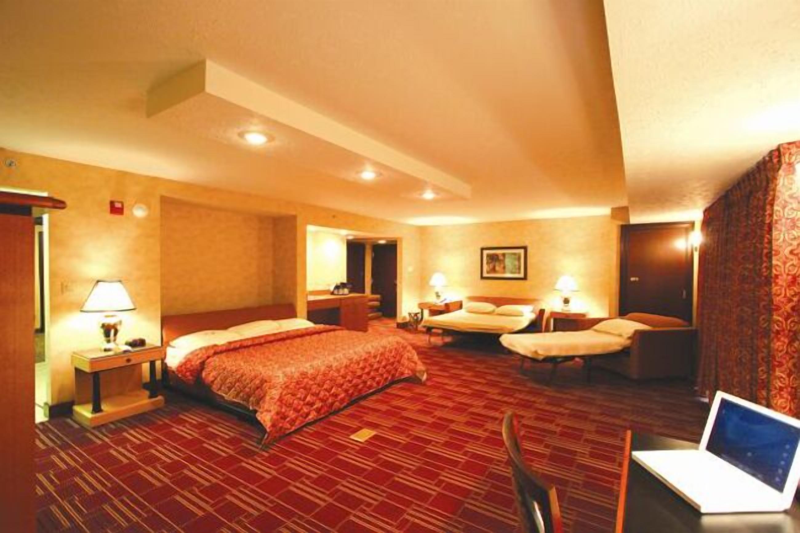 Habitación de hotel con dos camas, un escritorio con una computadora portátil y piso cubierto de alfombra roja.