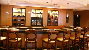 Una zona de bar con sillas y mesas de madera, una barra con vitrina y luces colgantes.