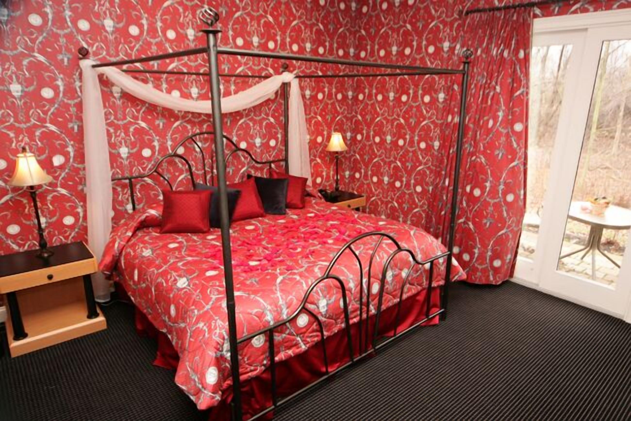 A four-poster bed with a red floral bedspread, two bedside lamps, and a view of the outdoors through a glass door.