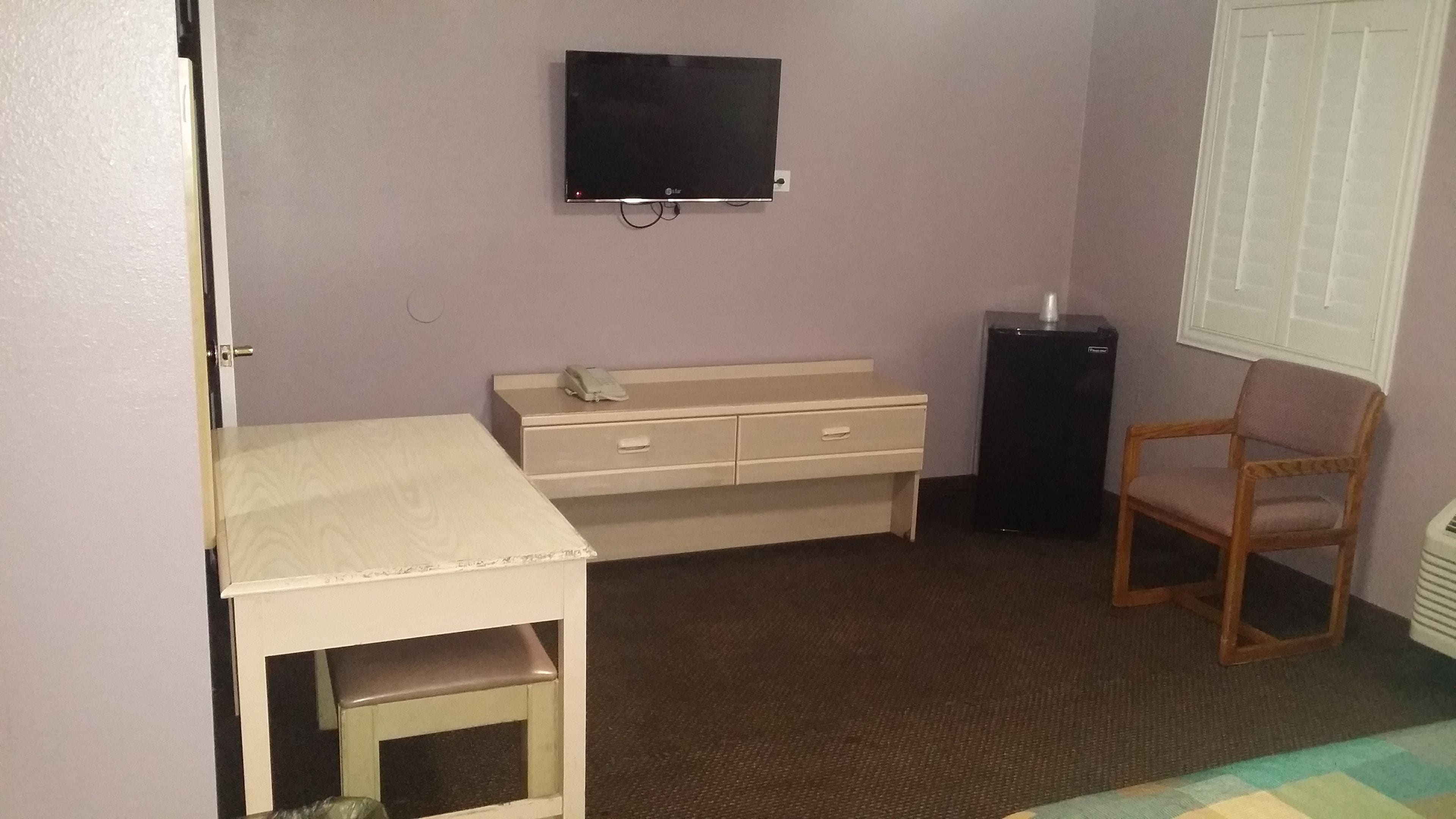 A small room with a desk, a chair, a television mounted on the wall, and a window with blinds.