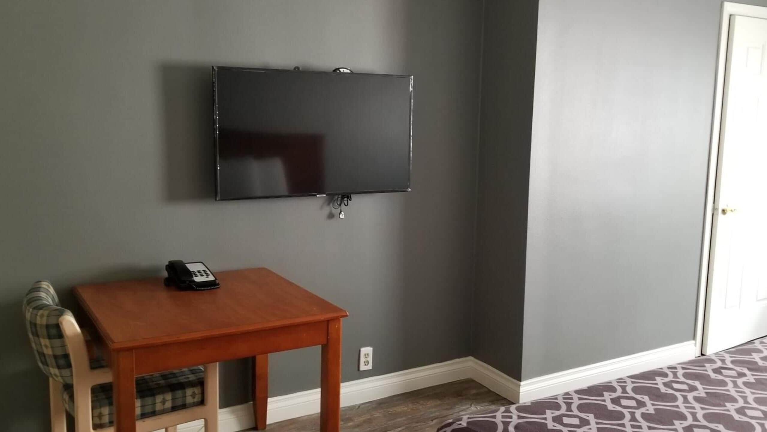 A room with a flat-screen TV mounted on the wall, a small wooden table with a telephone, and a plaid chair.
