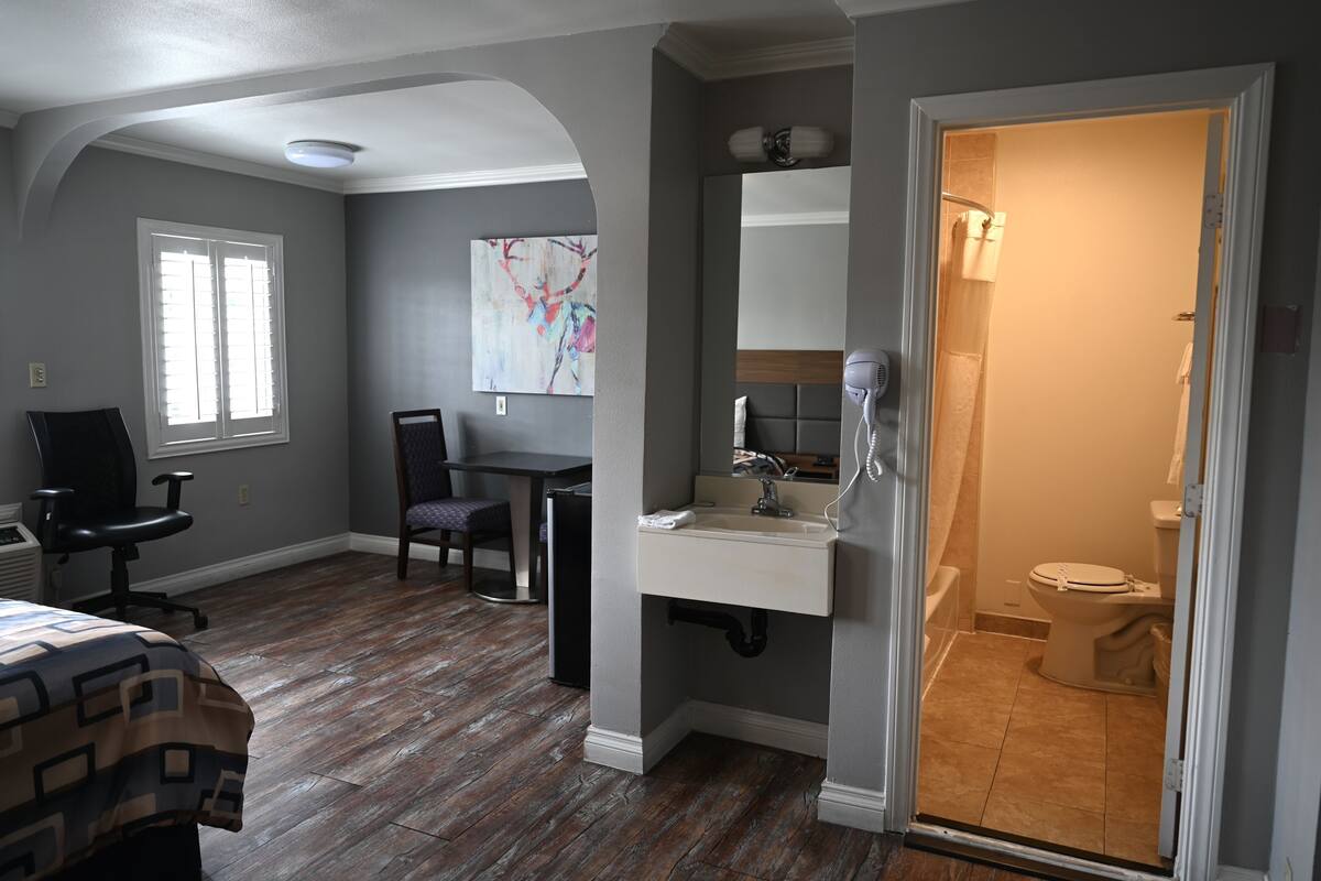 A room with a wooden floor, a desk with a chair, a bed, a bathroom with a toilet, and a shower.